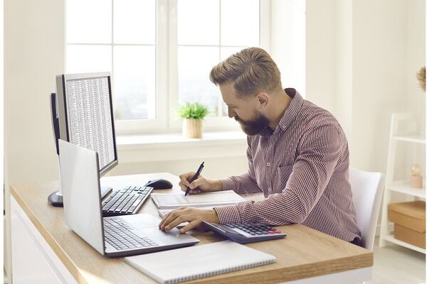 Person sitting at a small table writing notes beside a laptop and mug