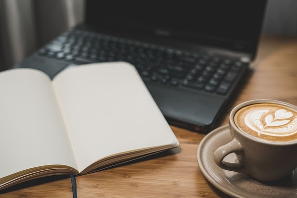Person sitting at a desk with a laptop, notebook, and warm drink in a bright office